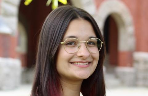 Sana smiles towards the camera in gold rimmed glasses and a maroon shirt. She has brown straight, long, hair with red lowlights. In the background a UNH brick building is visible. 